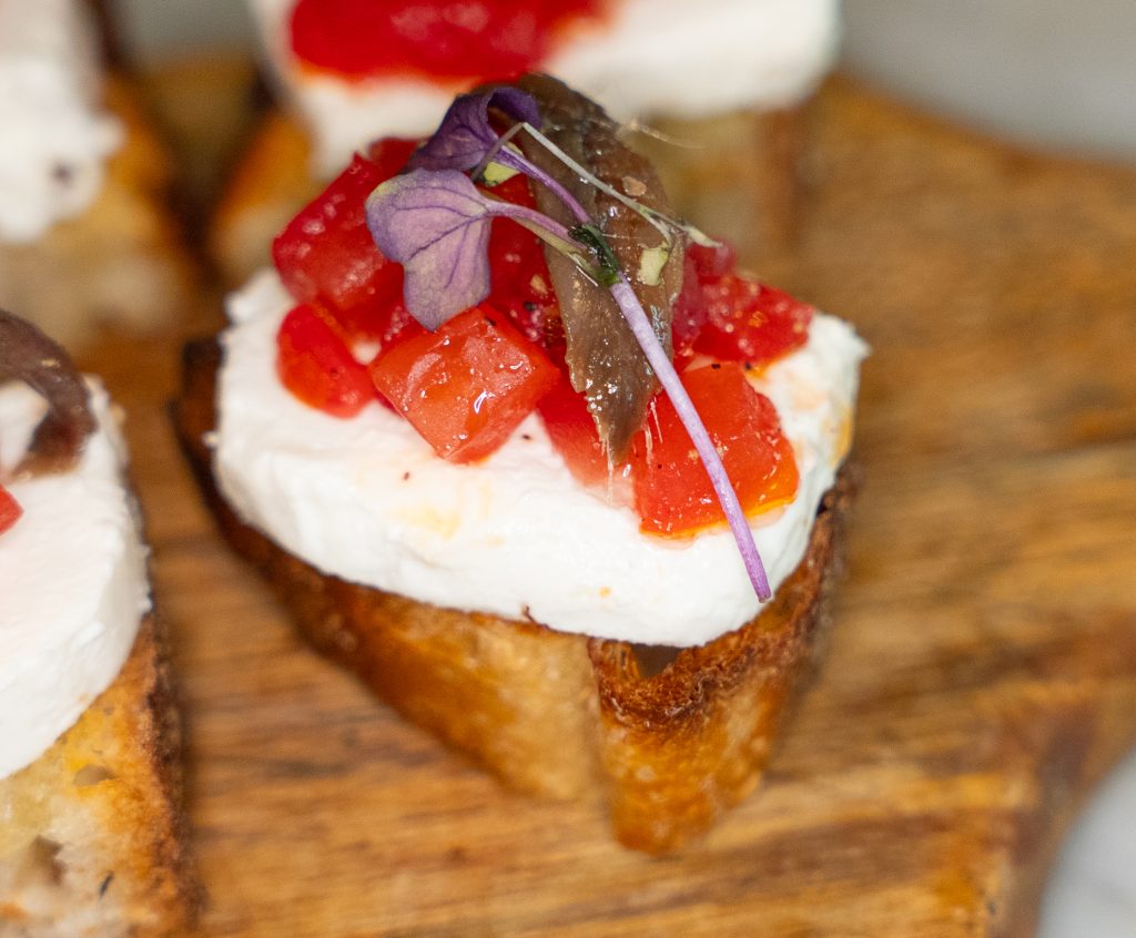 Close-up of gourmet bruschetta with creamy cheese, diced tomatoes, anchovy, and microgreens on toasted bread at a restaurant near Hudson Yards.