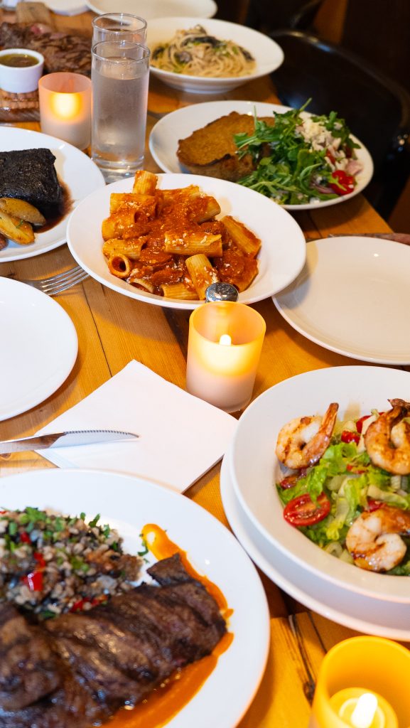 A table full of various Argentinian and grilled dishes including rigatoni pasta with tomato sauce, grilled steak, shrimp salad, short ribs, and assorted side dishes, surrounded by candles and water glasses.