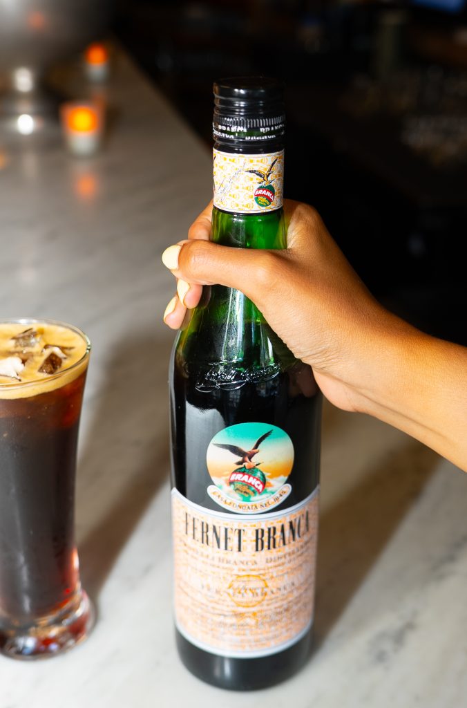 Hand holding a Fernet Branca bottle next to a cocktail with ice on a bar counter.