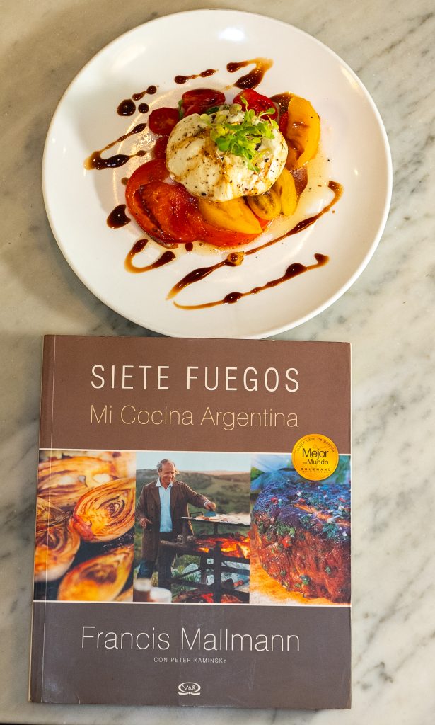 A tomato and burrata salad with balsamic glaze beside the Argentinian cookbook Siete Fuegos by Francis Mallmann, on a marble table near Hudson Yards.