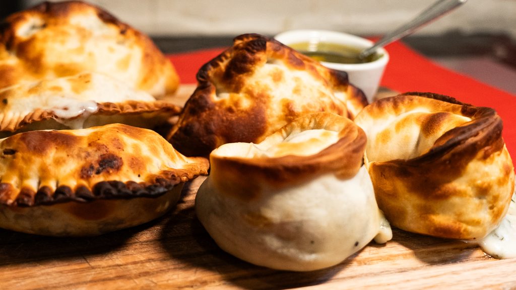 Freshly baked Argentinian empanadas served on a wooden board with chimichurri sauce.