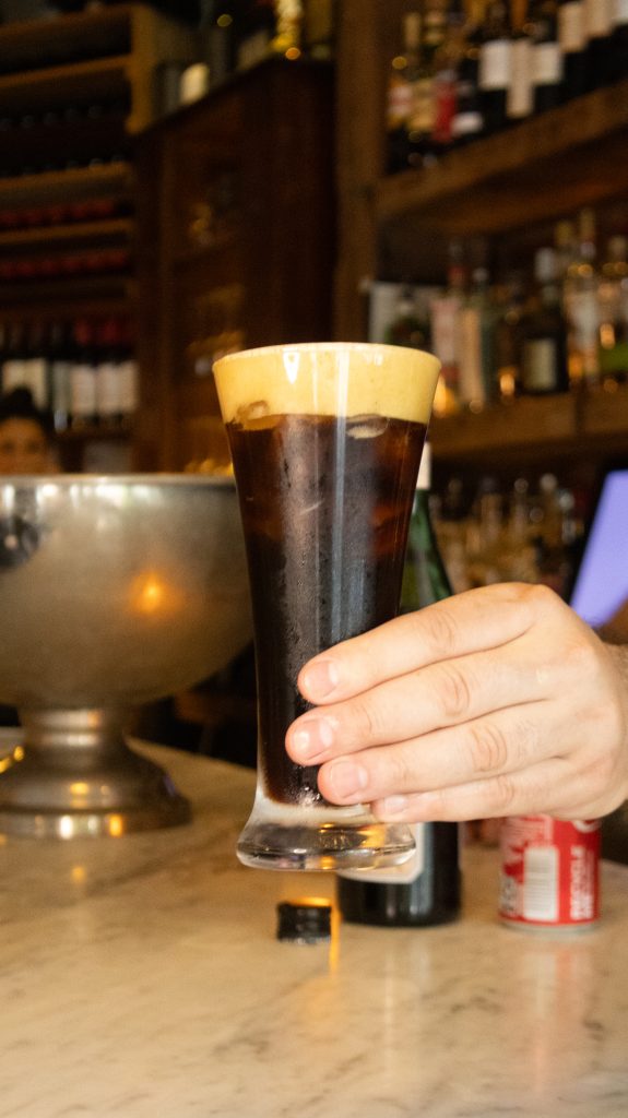 Hand holding a tall glass of Fernet Branca cocktail with foam on top, served at a bar counter.