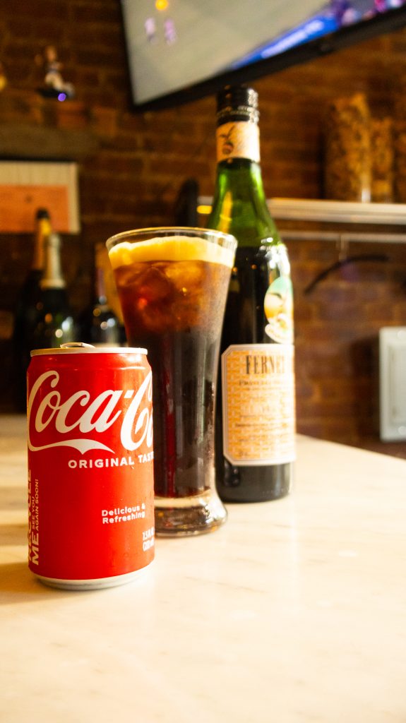 Bottle of Fernet Branca with a glass of Fernet and Coke, next to a Coca-Cola can on a wooden table.